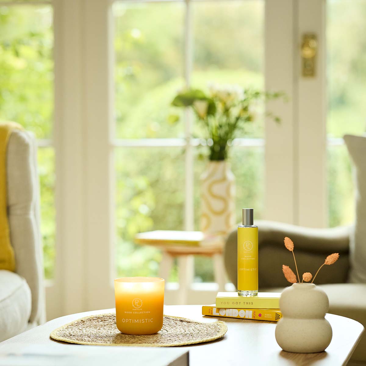 Optimistic Room Spray shown with the Optimistic Refillable Jar Candle on a table with windows and a vase of flowers in the background.