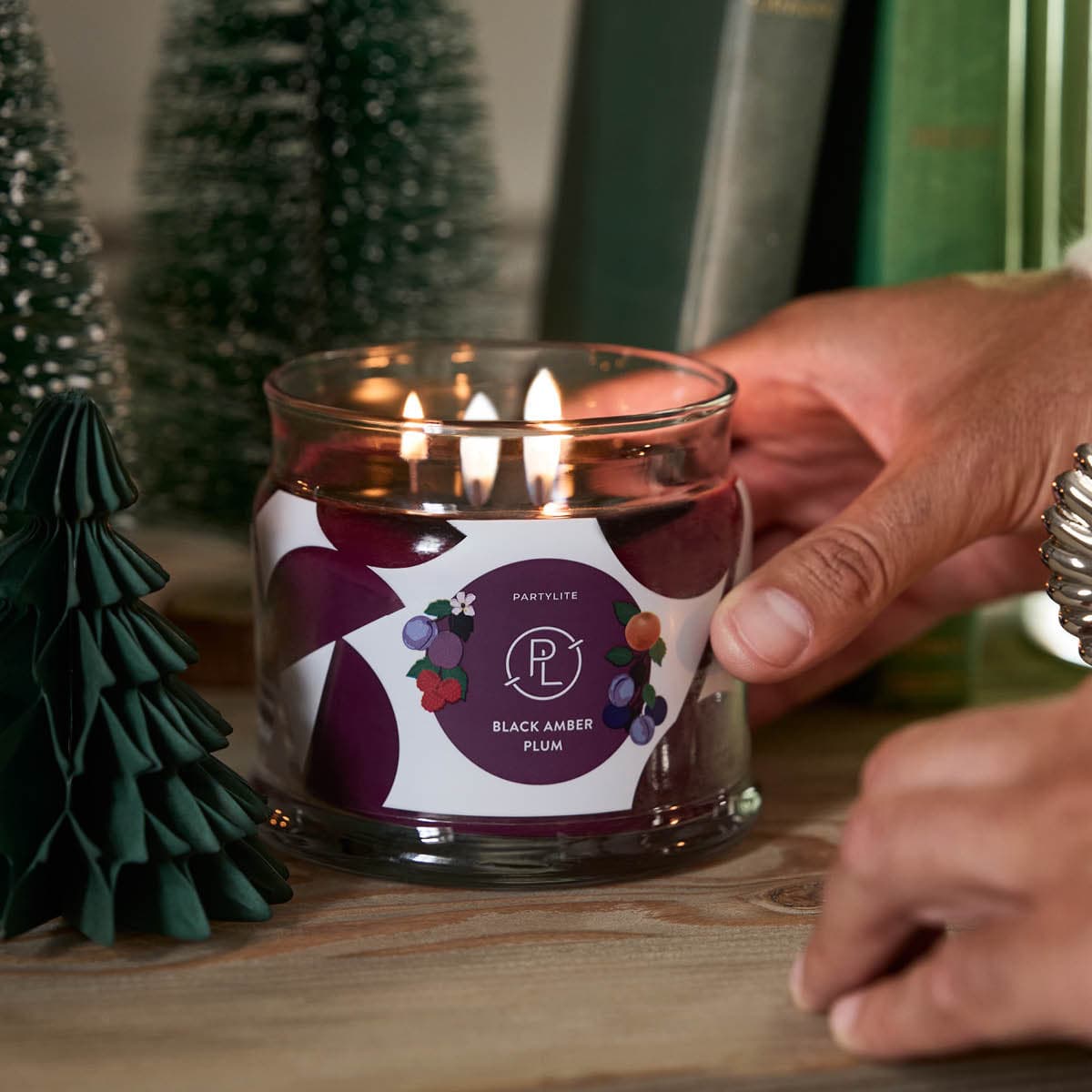 Black Amber Plum 3-Wick Jar Candle lit being put in place by a pair of hands on a wooden surface.
