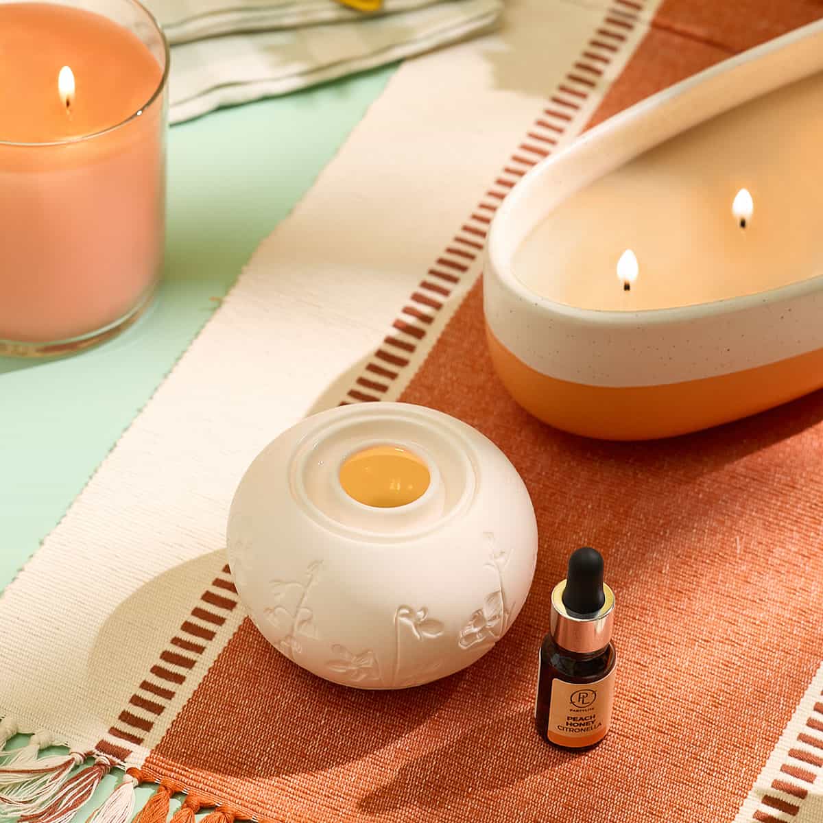 Peach Honey Citronella Fragrance Oil shown with a Fragrance Warmer and Peach Honey Citronella GloLite Jar and the two tone 4 wick Peach Honey Citronella jar on a woven table mat
